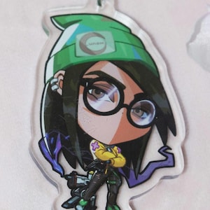 Könnte beinhalten: Ein grün-violetter Anime-Charakter-Schlüsselanhänger mit grüner Mütze und Brille. Der Schlüsselanhänger hat ein weißes Logo mit der Aufschrift "VGN" auf der Mütze.