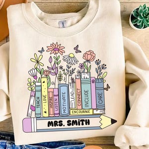 May include: Cream-colored sweatshirt featuring a colorful floral and book design. The design includes a row of books with words like "TEACH," "LOVE," and "INSPIRE." A pencil with "MRS. SMITH" is at the bottom. Butterflies and flowers are also included.