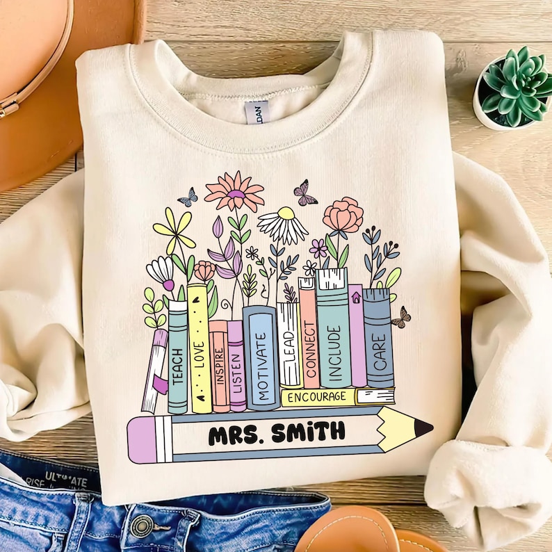 May include: Cream-colored sweatshirt featuring a colorful floral and book design. The design includes a row of books with words like "TEACH," "LOVE," and "INSPIRE." A pencil with "MRS. SMITH" is at the bottom. Butterflies and flowers are also included.
