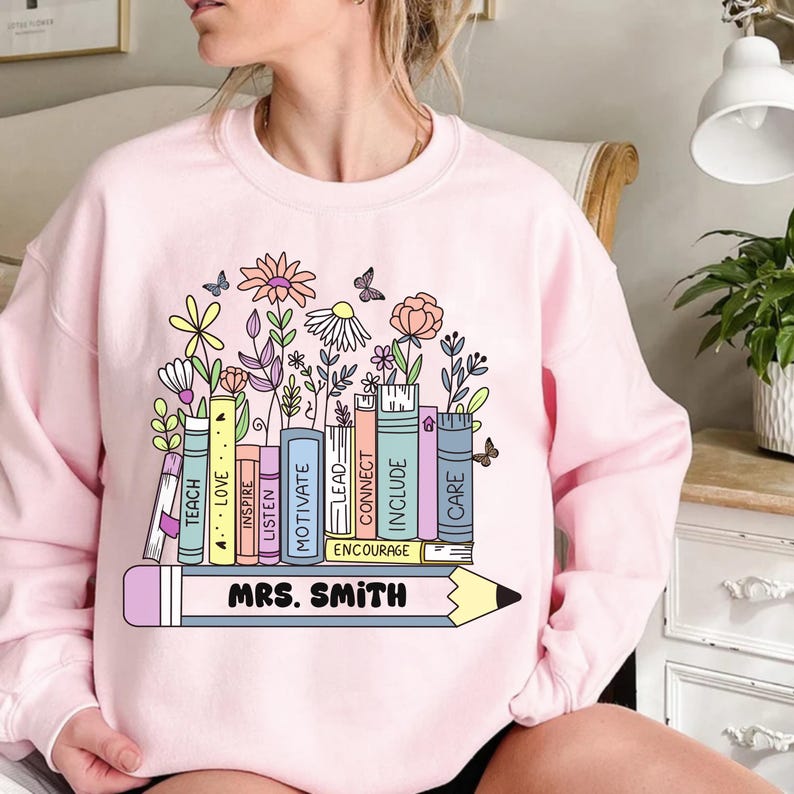 May include: A light pink sweatshirt featuring a colorful graphic. The design includes a row of books with words like "TEACH" and "CARE," topped with flowers and butterflies. A large pencil with "MRS. SMITH" is at the bottom.