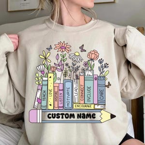 May include: Beige sweatshirt featuring a colorful illustration of books with floral accents and butterflies. The books have words like "TEACH", "LOVE", and "MOTIVATE". A pencil with "CUSTOM NAME" is at the bottom. A charming and educational design.