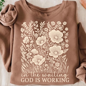May include: A brown sweatshirt with a cream floral design. The design features large flowers and foliage, with the text "in the waiting GOD IS WORKING" at the bottom. The sweatshirt has a crew neck and long sleeves.