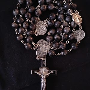 May include: A rosary with dark gray, speckled beads and silver-toned metal accents. The rosary features a crucifix with a figure of Jesus and several round medallions with religious symbols.