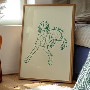 May include: A framed wall art print featuring a green line drawing of a spotted dog on a cream background. The dog is depicted in a reclining position with the word BANDIT written in green capital letters above its back.