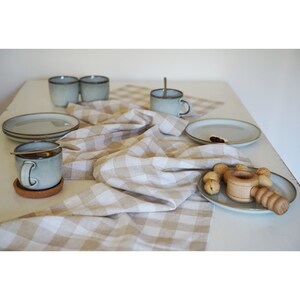 Pode incluir: Um corredor de mesa de linho xadrez bege e branco com uma caneca de cerâmica azul claro, um prato com nozes e um quebra-nozes de madeira em uma mesa branca.
