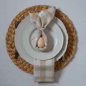 May include: A white plate with a brown and white gingham napkin folded into a bunny shape and an egg inside. The plate is on a woven placemat.