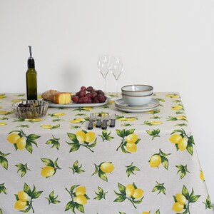 May include: A white tablecloth with a lemon print pattern. The tablecloth is set with a bowl of olives, a plate of grapes, a plate of cheese and bread, two wine glasses, and a bowl.