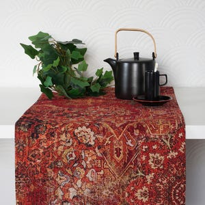 Peut inclure: Un chemin de table rectangulaire orné d'un motif floral complexe dans des tons rouge profond, orange et noir. Il est présenté sur une surface blanche avec une théière noire, une tasse assortie sur sa soucoupe et une plante verte.
