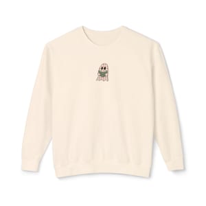 May include: A cream-colored crewneck sweatshirt featuring a small, centered embroidered graphic of a ghost with a pink and white checkered pattern, holding a small green book. The ghost has two dark oval eyes and a simple outline design.