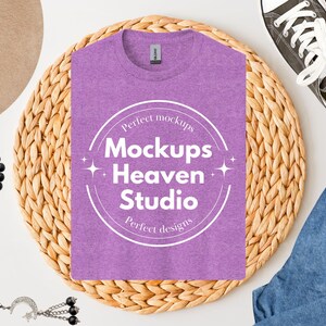 May include: A heather purple t-shirt with the text "Mockups Heaven Studio" in a white circular design. The shirt is on a woven mat, with a pair of canvas sneakers, blue jeans, a hat, and a beaded necklace.