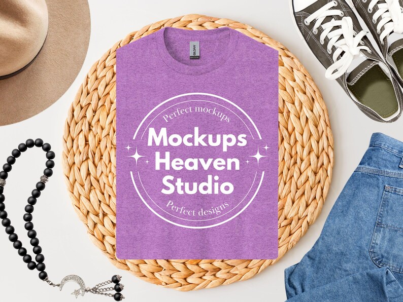 May include: A heather purple t-shirt with the text "Mockups Heaven Studio" in a white circular design. The shirt is on a woven mat, with a pair of canvas sneakers, blue jeans, a hat, and a beaded necklace.