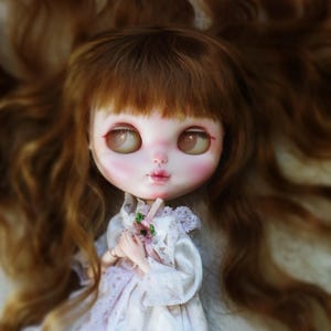 Könnte beinhalten: Eine Puppe mit hellem Teint, großen Augen und einem süßen Ausdruck. Die Puppe hat langes, welliges, rotbraunes Haar und trägt ein weißes Kleid mit Spitzendetails. Sie hält einen kleinen Strauß rosa Blumen. Das Gesicht der Puppe ist zart mit rosigen Wangen und Lippen bemalt.