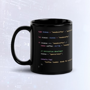 Può includere: Una tazza in ceramica nera con codice di programmazione colorato stampato sul lato. Il codice include righe come type Status = "needsCoffee", const coffee: string = "intoMug" e console.log("Coffee loaded. Ready for production").