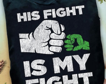 May include: Black t-shirt featuring the text "HIS FIGHT IS MY FIGHT" in white, accompanied by a graphic of two fists, one white and one green. The t-shirt has a crew neck.