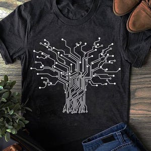 May include: Dark gray t-shirt featuring a white graphic of a tree composed of circuit board lines and nodes. The design resembles a digital tree, with branches and a trunk made of interconnected lines. The shirt is displayed on a wooden surface.