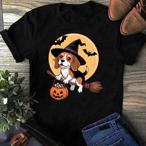 Könnte beinhalten: Schwarzes T-Shirt mit einem Cartoon-Beagle, der als Hexe verkleidet ist, einen schwarzen Hut mit lila Band trägt und einen Kürbis-Eimer voller Süßigkeiten hält. Der Beagle reitet auf einem Besen vor einem Vollmond mit Fledermäusen.