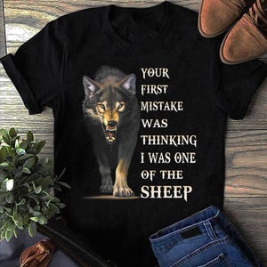 May include: Black t-shirt featuring a snarling wolf illustration with the text "YOUR FIRST MISTAKE WAS THINKING I WAS ONE OF THE SHEEP." The wolf is depicted with detailed fur and glowing eyes, creating a fierce aesthetic.