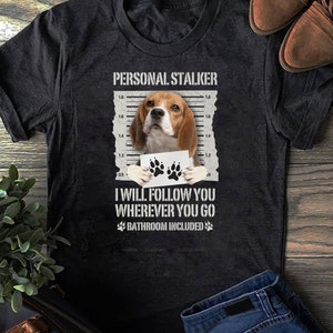 Könnte beinhalten: Ein schwarzes T-Shirt mit einer weißen Grafik eines Beagle-Hundes, der ein Schild mit Pfotenabdrücken hält. Der Text "PERSONAL STALKER" befindet sich über dem Hund und "I WILL FOLLOW YOU WHEREVER YOU GO BATHROOM INCLUDED" unter dem Hund.