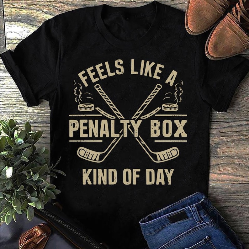 Op de afbeelding: Zwart T-shirt met een hockeystick-afbeelding en de tekst "Feels like a penalty box kind of day".