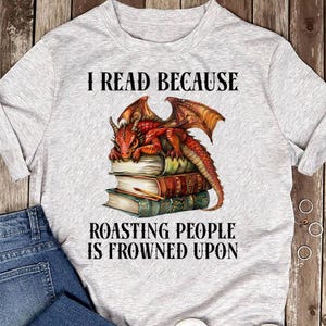 May include: Light gray t-shirt with the text "I READ BECAUSE ROASTING PEOPLE IS FROWNED UPON." The shirt features a red dragon with orange wings resting on a stack of books. The books have brown, green, and cream-colored covers.
