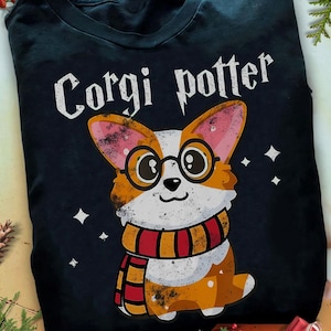 Puede incluir: Camiseta azul marino con un corgi de dibujos animados con gafas y una bufanda. El texto "Corgi Potter" está impreso encima de la ilustración del corgi. El corgi es naranja, blanco y negro con una bufanda roja y dorada.