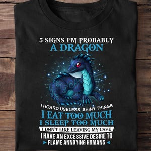Może przedstawiać: Czarny T-shirt z ilustracją niebieskiego smoka i tekstem "5 Signs I'm Probably A Dragon". Tekst pod smokiem brzmi: "I hoard useless, shiny things I eat too much I sleep too much I don't like leaving my cave I have an excessive desire to flame annoying humans."