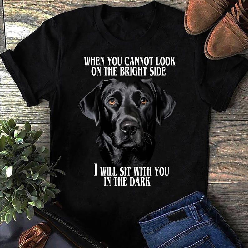 May include: Black t-shirt featuring a black Labrador Retriever portrait with the text "WHEN YOU CANNOT LOOK ON THE BRIGHT SIDE, I WILL SIT WITH YOU IN THE DARK." The shirt is paired with blue jeans and brown shoes.