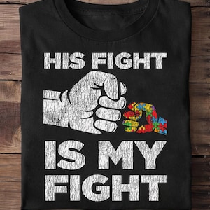 Puede incluir: Camiseta negra con el texto blanco "HIS FIGHT IS MY FIGHT" sobre un gráfico de dos puños. Un puño es blanco, el otro es un diseño de rompecabezas colorido, que simboliza la concienciación sobre el autismo.
