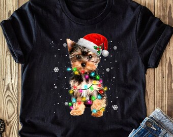 Może przedstawiać: Czarny T-shirt z Yorkshire Terrierem w czapce Świętego Mikołaja i światełkami choinkowymi. Pies jest otoczony przez płatki śniegu.