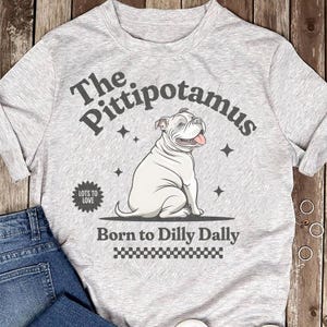 Pode incluir: Camiseta cinza claro com uma ilustração de um buldogue branco sentado. O texto na camiseta diz "The Pittipotamus" acima do cão e "Born to Dilly Dally" abaixo. Há também um pequeno emblema "Lots to Love".