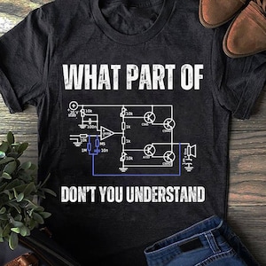 Op de afbeelding: Zwart t-shirt met witte tekst en een elektrisch schema. De tekst luidt "WHAT PART OF DON'T YOU UNDERSTAND". Het schema bevat verschillende elektronische componenten en labels. Bruine schoenen en een blauwe jeans zijn zichtbaar.