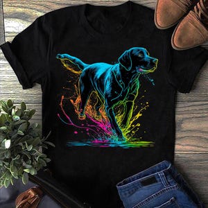 May include: Black t-shirt with a neon-style illustration of a dog. The dog is outlined in blue and black, with colourful splashes of pink, yellow, and green. The design is set against a black background, creating a striking contrast.