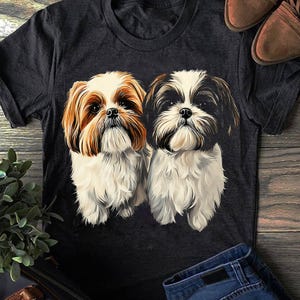 Op de afbeelding: Donkergrijs t-shirt met een gedetailleerde illustratie van twee Shih Tzu honden. Eén hond heeft bruin en wit haar, de andere zwart en wit. De honden staan centraal op het shirt, met een houten oppervlak en jeans op de achtergrond.