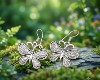 May include: A pair of silver-toned metal earrings shaped like butterflies with textured, dotted wing patterns. Each earring features a curved hook wire for attachment. The text "The Unique Gift Art" is visible in the bottom right corner.