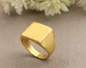 Könnte beinhalten: Ein goldfarbener Siegelring mit einer glatten, polierten quadratischen Oberfläche und einer massiven, sich verjüngenden Ringschiene. Der Ring hat ein reflektierendes metallisches Finish und ein klares, geometrisches Design.
