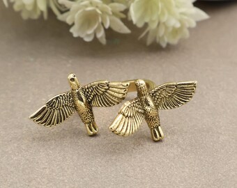 May include: A gold-toned metal ring featuring two birds with outstretched wings. The birds are positioned side-by-side, facing opposite directions, with detailed feather textures visible on their wings and bodies. The band connects the two birds.