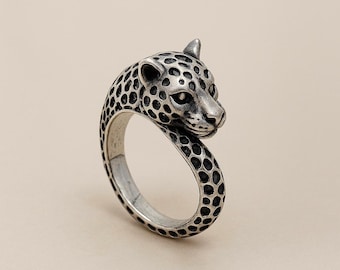 May include: A silver-toned metal ring featuring a sculpted leopard head design. The band and the feline head are covered in a detailed, dark-oxidised spotted pattern that mimics the natural markings of a leopard, creating a textured appearance.