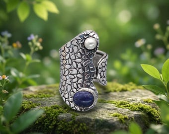 May include: A wide silver ring featuring a textured, pebble-like surface pattern. The ring is adorned with a single small white pearl at the top and a larger, smooth oval-shaped blue stone at the bottom, creating a distinct contrast in shape and color.