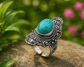 May include: An oval turquoise gemstone set in a silver-toned metal ring. The metal band features intricate, raised scrollwork and filigree patterns surrounding the central stone. The ring has an open, adjustable band design at the bottom.