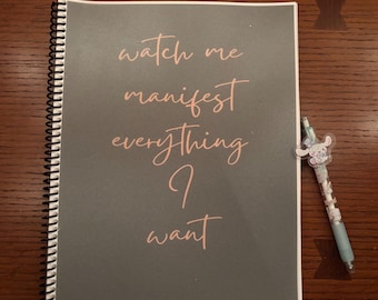 May include: A spiral-bound notebook with a gray cover featuring the text "watch me manifest everything I want" in pink script. A light blue pen with a cartoon character charm rests on the right side of the notebook.