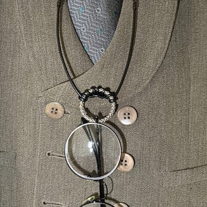 以下が含まれることがあります： 黒いフレームの丸い眼鏡が、銀色のリングが付いた黒いコードネックレスから吊り下げられています。 ネックレスは、木製のボタンが付いた茶色のスーツジャケットの上にかけられています。 柄物のネクタイが見えます。