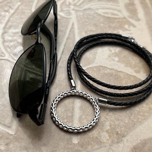 Op de afbeelding: Zwarte zonnebril met donkere glazen ligt naast een zwarte leren ketting. De ketting heeft een zilverkleurige, open cirkel hanger met een textuurontwerp. De ketting is gevlochten en heeft een zilveren sluiting.