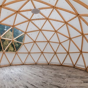 May include: A wooden geodesic dome structure with a white fabric covering. The dome has a large window that looks out onto a forest. The interior of the dome has a wooden floor.