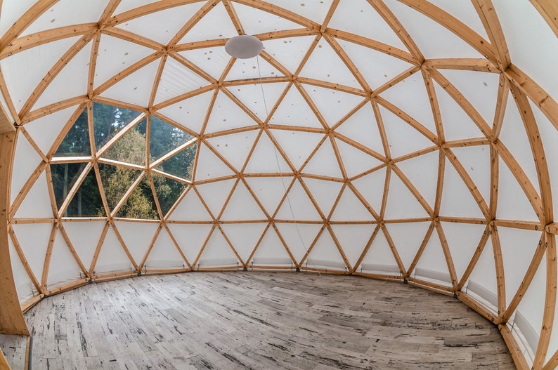 May include: A wooden geodesic dome structure with a white fabric covering. The dome has a large window that looks out onto a forest. The interior of the dome has a wooden floor.
