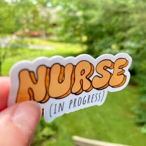 May include: A white sticker with the word "NURSE" in orange bubble letters. Below the word is the text "(IN PROGRESS)" in black.