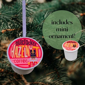 May include: A white coffee pod ornament with a pink and yellow design that says "Nutcracker Hazelnut Coffee". The ornament is hanging from a white string on a green Christmas tree.