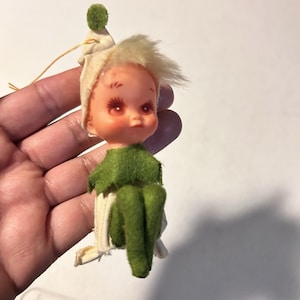 Op de afbeelding: Een kleine elfenpop als ornament met een bleek gezicht, roze wangen en blond haar. De pop draagt een crèmekleurige puntmuts met een groene pompon, een groene vilten kraag en groene vilten benen. Een dun gouden koordje zit aan de muts vast.