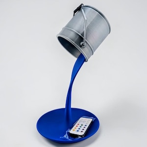 May include: A unique desk lamp featuring a gray bucket seemingly pouring blue paint. The paint flows down to form the lamp's stand and base. A remote control sits on the base. The lamp is a creative home decor item.