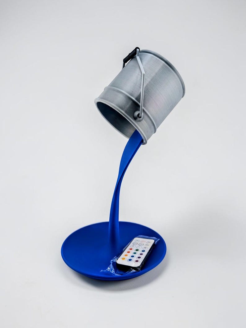 May include: A unique desk lamp featuring a gray bucket seemingly pouring blue paint. The paint flows down to form the lamp's stand and base. A remote control sits on the base. The lamp is a creative home decor item.