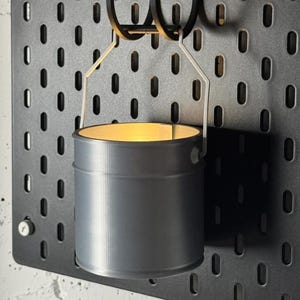 May include: A gray metal bucket-shaped candle holder suspended from a black pegboard. The candle holder has a cylindrical shape with a light source inside, casting a warm glow. The pegboard has evenly spaced oval holes.